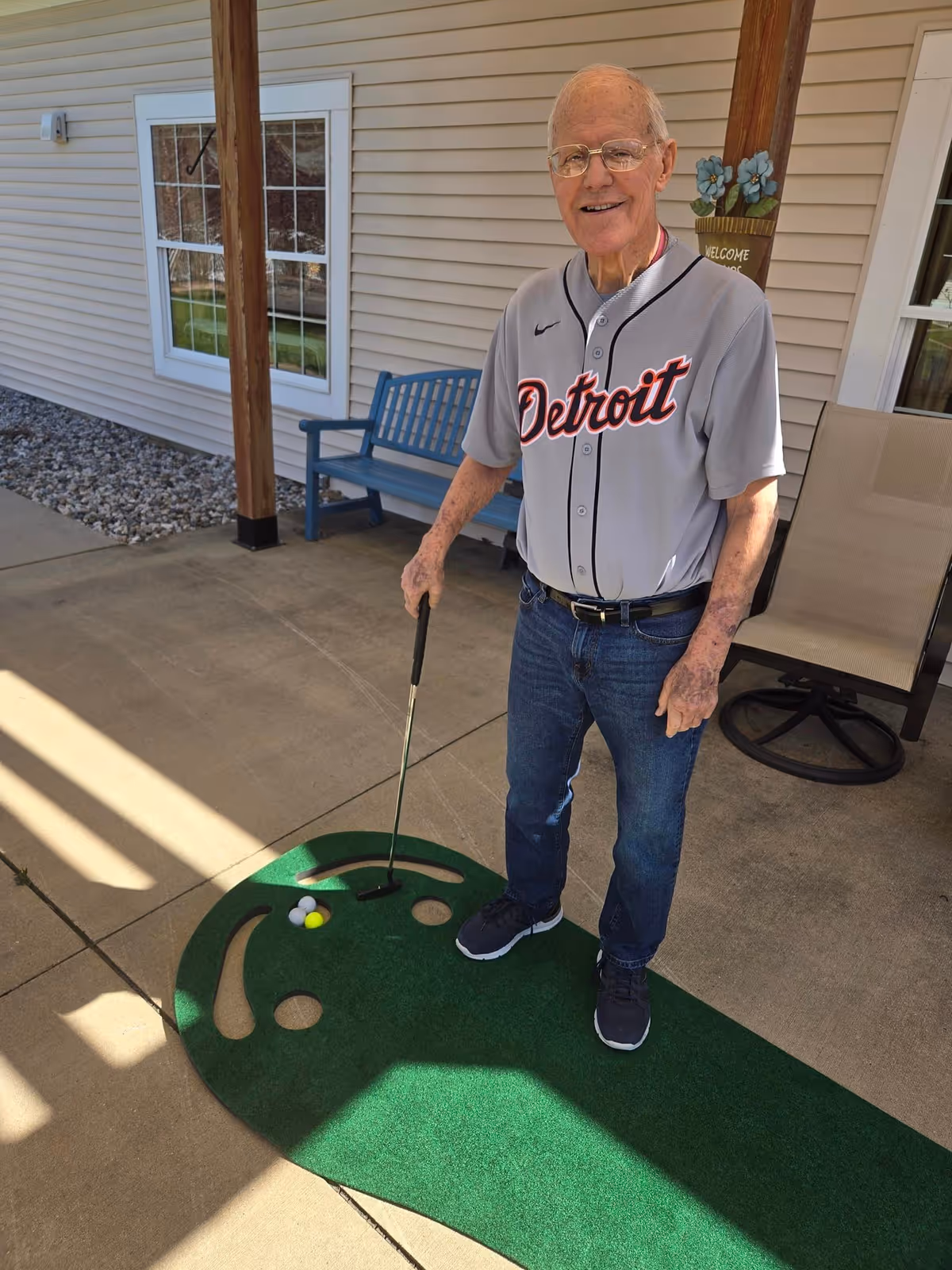An elderly man wearing a Detroit baseball jersey and jeans is standing on a concrete patio next to a small putting green mat with golf balls. He is holding a golf putter and smiling. Behind him is a blue bench, a beige chair, and a house wall with windows and a welcome sign with flowers.