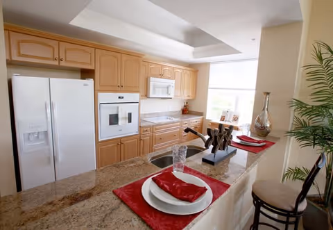 Bright kitchen with light wood cabinets, white refrigerator, built-in oven, microwave, and a granite countertop island set with two place settings featuring red napkins and placemats. A decorative sculpture and a glass of water are on the island, with a tall plant and a window in the background.
