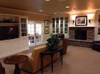 A cozy living room with beige walls and carpet, featuring a stone fireplace with a framed picture above it, built-in white cabinets with glass doors, a flat-screen TV, a brown leather chair, a tan sofa with a black console table behind it holding a plant, and a round wooden side table.
