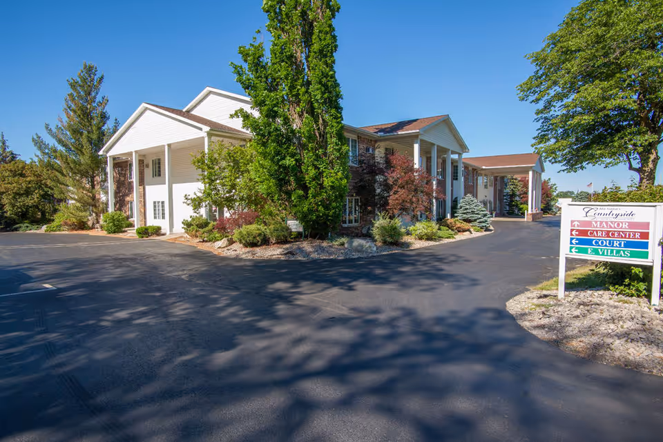 Exterior view of Countryside Manor, a two-story building with white columns and surrounded by trees and shrubs, under a clear blue sky. A paved driveway curves around the building, and a signpost indicates directions to Manor, Care Center, Court, and E. Villas.