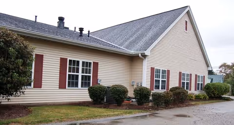 Exterior view of a single-story building with beige siding and a gray shingled roof. The building has multiple windows with red shutters and is surrounded by small bushes and a paved area.