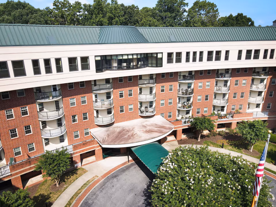 Exterior view of a multi-story senior living facility with a green metal roof, red brick facade, and multiple balconies. There is a covered entrance with a curved canopy and a driveway in front. Trees and landscaping surround the building, and an American flag is visible on the right side.