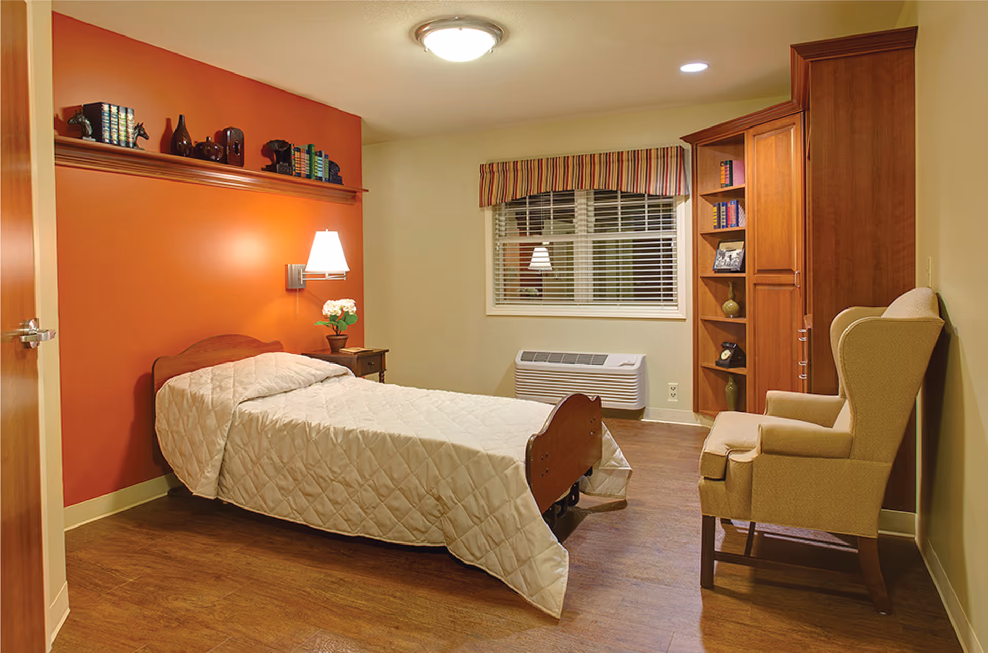 A cozy bedroom with a single bed covered in a white quilted bedspread, a wooden headboard, and a small bedside table with a lamp and a potted plant. The room has an orange accent wall with a shelf holding books and decorative items. There is a window with striped valance, a beige armchair, and a wooden cabinet with shelves containing books and decorative objects. The floor is wooden.