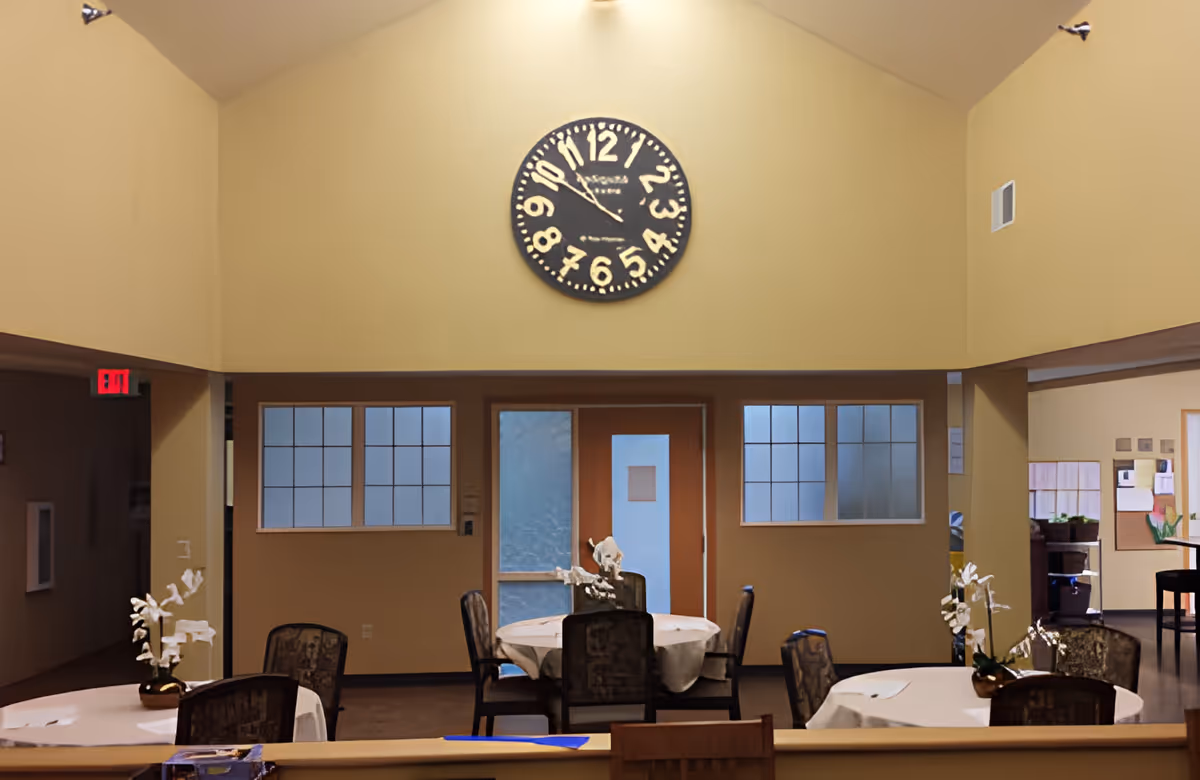 Interior communal dining area with round tables and chairs, floral centerpieces, and a large clock on the high wall.