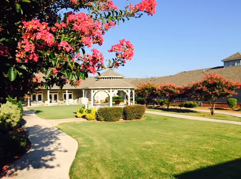 A sunny outdoor garden area at Castle Rock Assisted Living featuring a well-maintained lawn, a white gazebo in the center, flowering trees with bright pink blossoms, and a paved walkway curving through the grass. The background shows a single-story building with a porch and seating area.