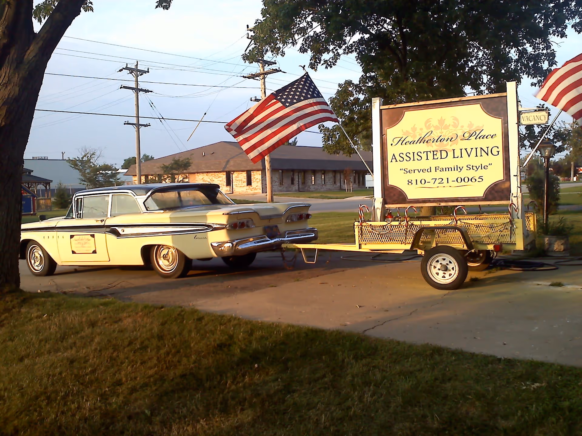A vintage car parked beside a trailer-mounted sign for Heatherton Place Assisted Living with American flags in front of the building.