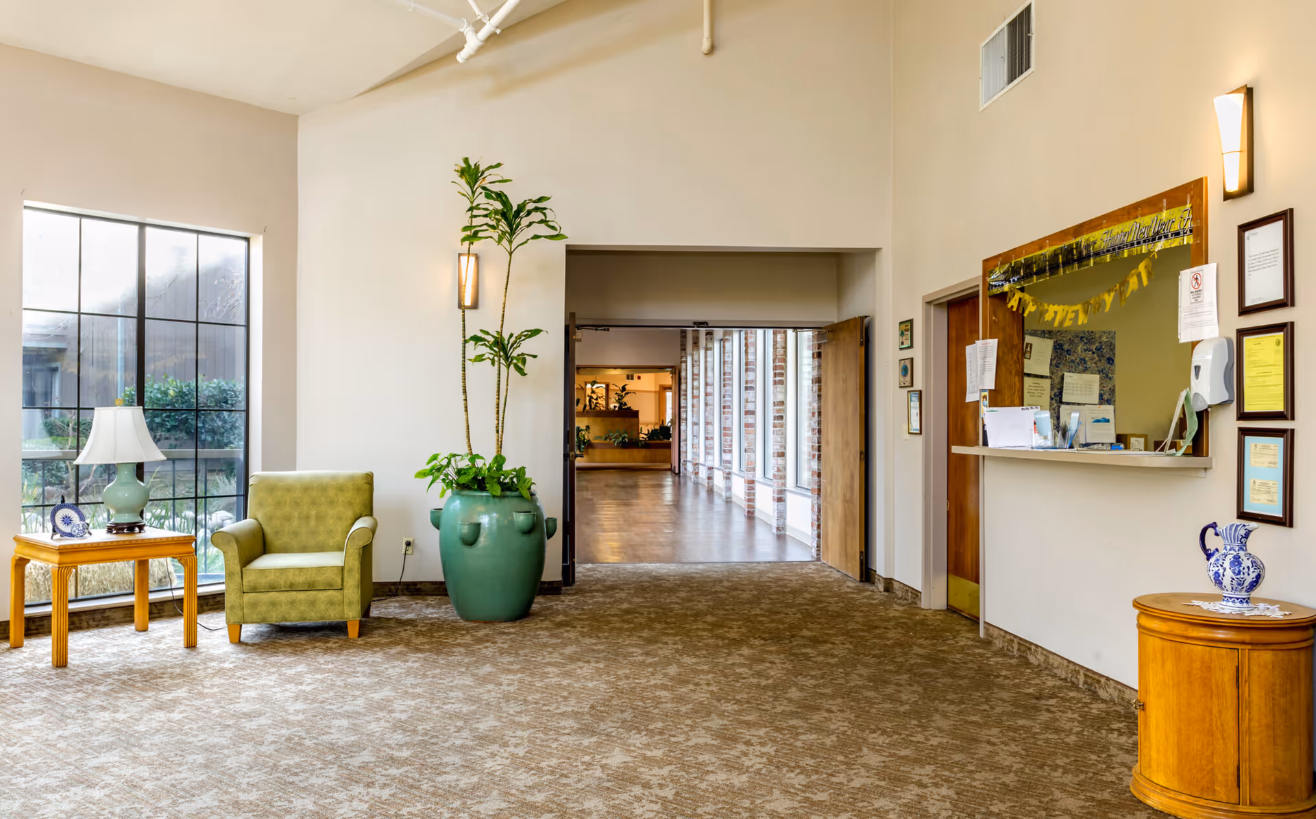 A bright and spacious interior area of The Atrium Of Carmichael featuring a green armchair next to a wooden side table with a lamp and decorative plate. A large green potted plant is placed near an open doorway leading to a hallway with large windows and brick walls. On the right side, there is a reception window decorated with a Happy New Year banner and various notices, along with a small wooden cabinet holding a blue and white ceramic pitcher.