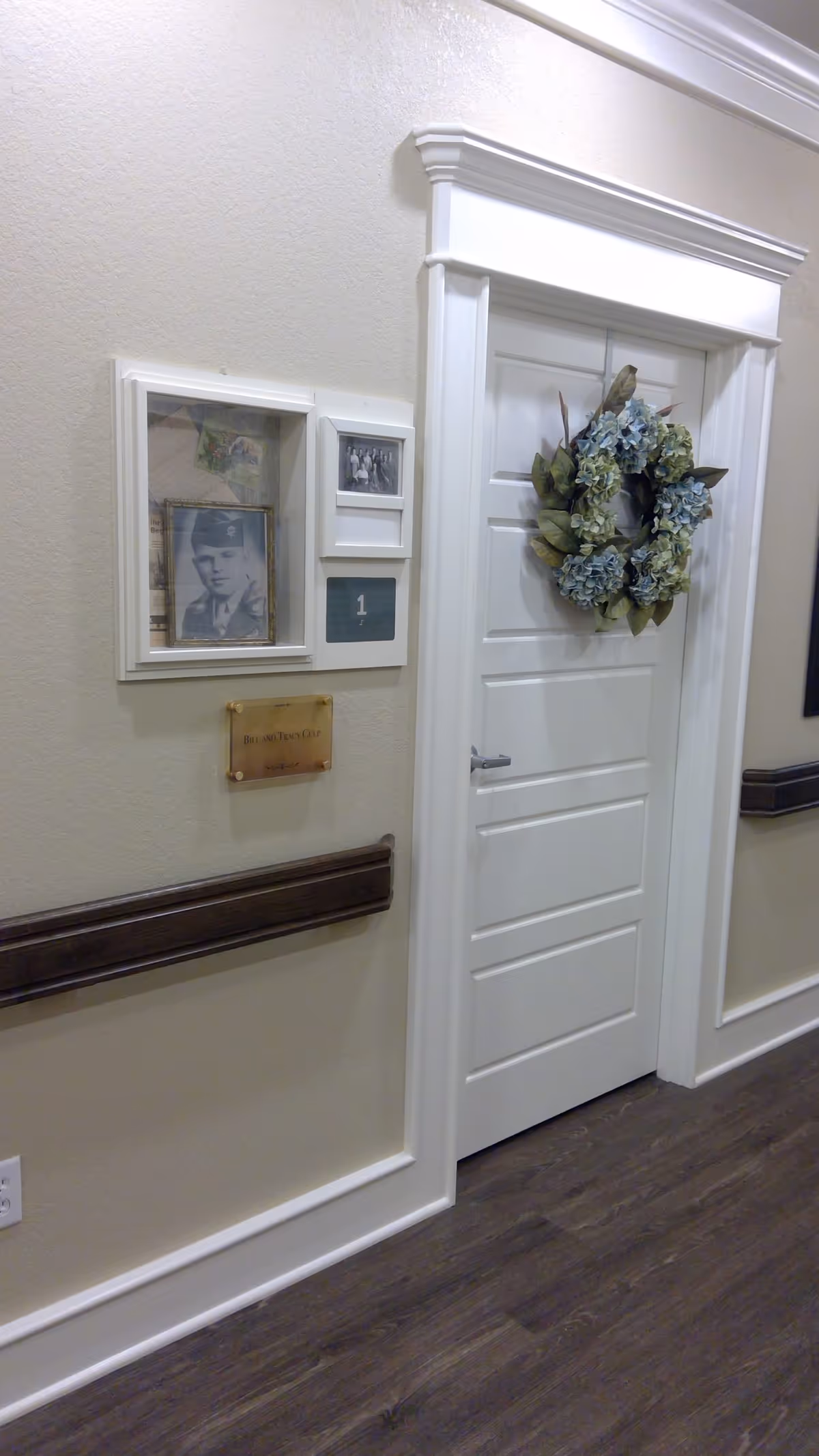 A white door decorated with a wreath made of green and blue flowers in a hallway with beige walls and dark wood flooring. Next to the door is a display case with framed photographs and a plaque that reads 'Bill and Tracy Cole'. The door has a sign with the number 1 on it.