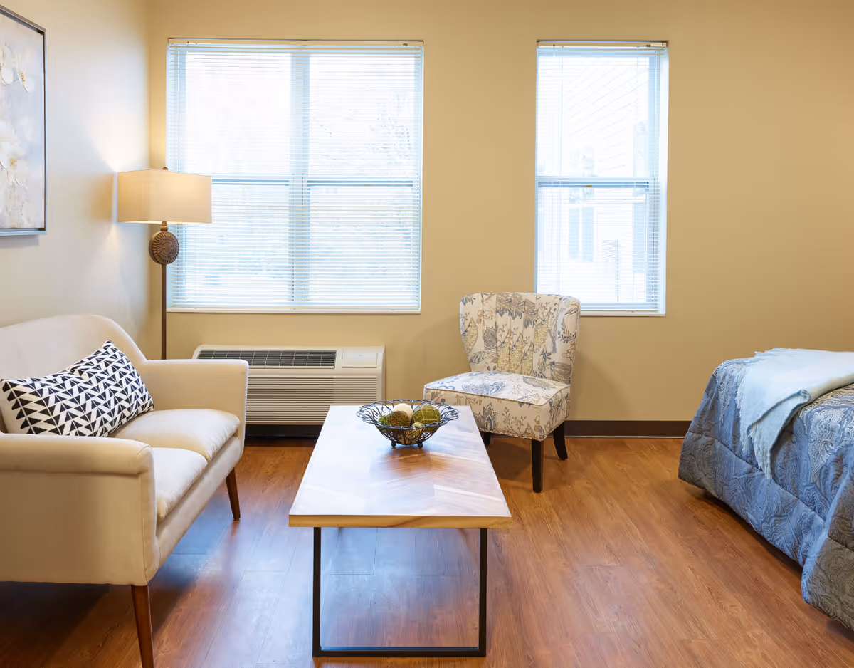 A bright senior living bedroom with a small seating area including a loveseat, patterned armchair, coffee table, and a bed by two windows.