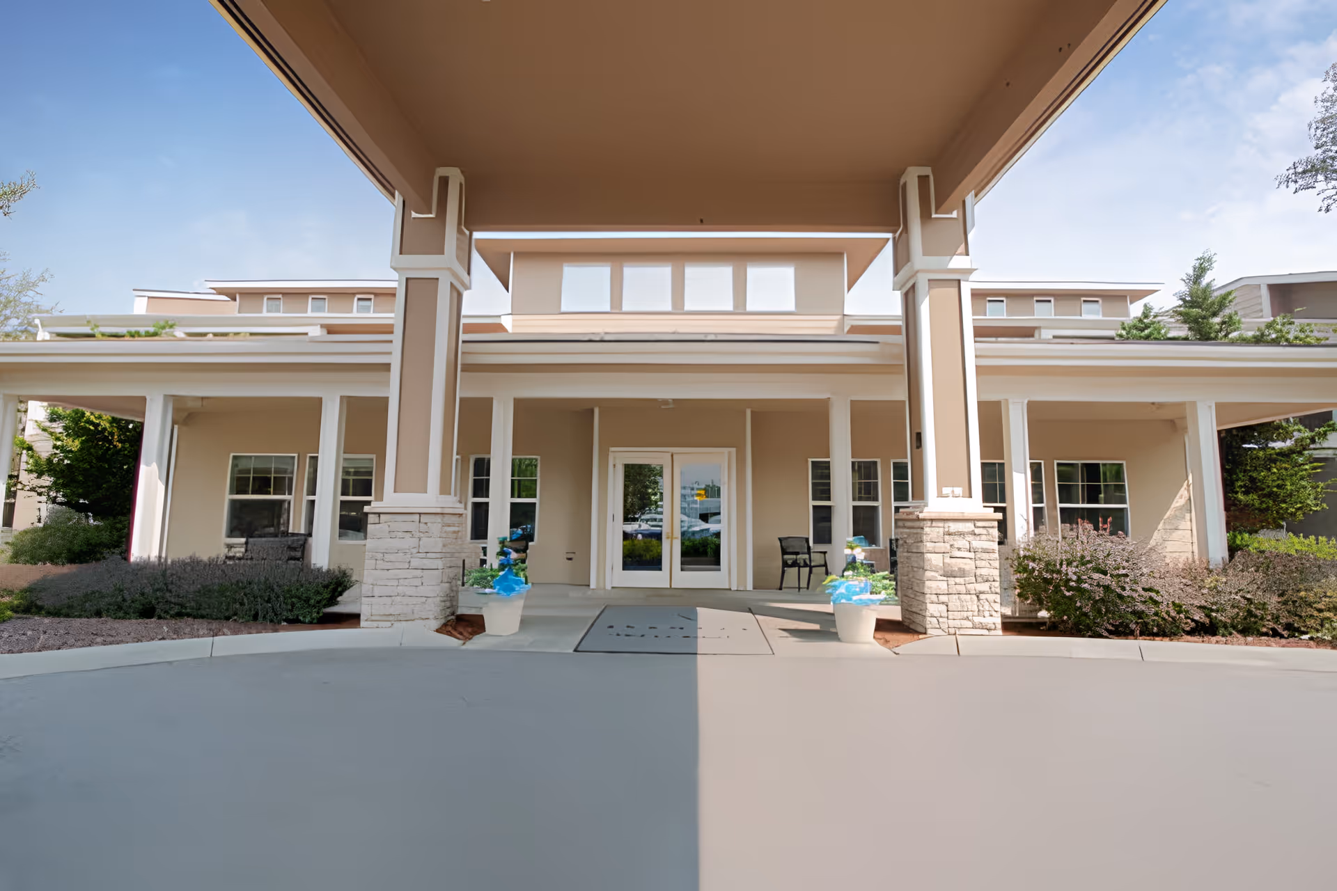 Covered porte-cochere and entrance of a beige senior living building with columns, planters, and double glass doors.