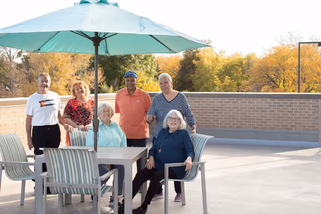 A group of six elderly people, three men and three women, are gathered on an outdoor patio. Two women are seated at a table with striped chairs under a large blue umbrella, while the others stand behind them. The background shows a brick wall and trees with autumn foliage.