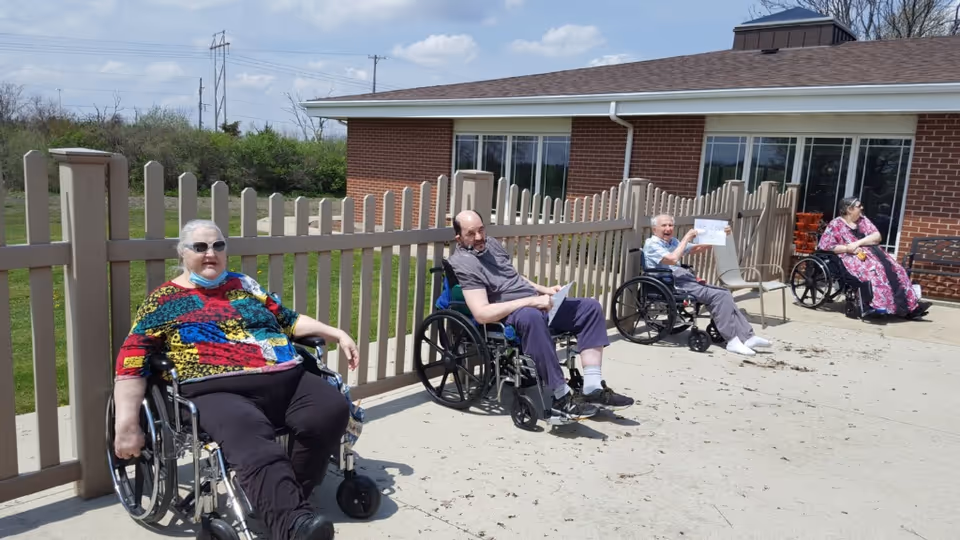 Four elderly individuals in wheelchairs sitting outside on a sunny day near a beige fence and a brick building. They appear to be enjoying the outdoor space with greenery in the background under a partly cloudy sky.