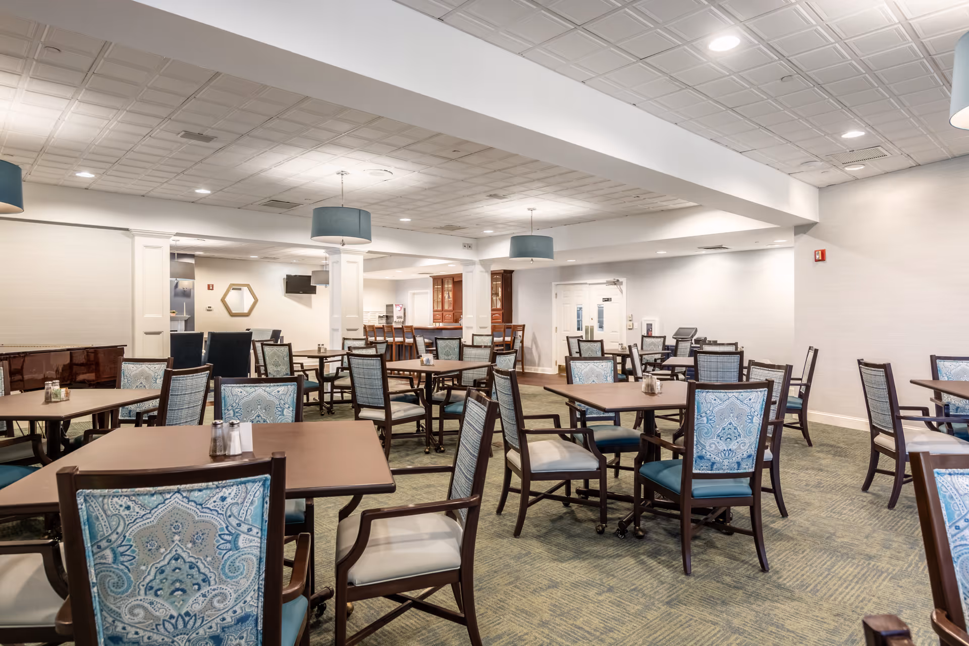A spacious dining room with multiple wooden tables and chairs featuring blue patterned upholstery. The room has a light-colored ceiling with recessed lighting and hanging blue lampshades. There is a piano on the left side and a bar area with high chairs in the background.