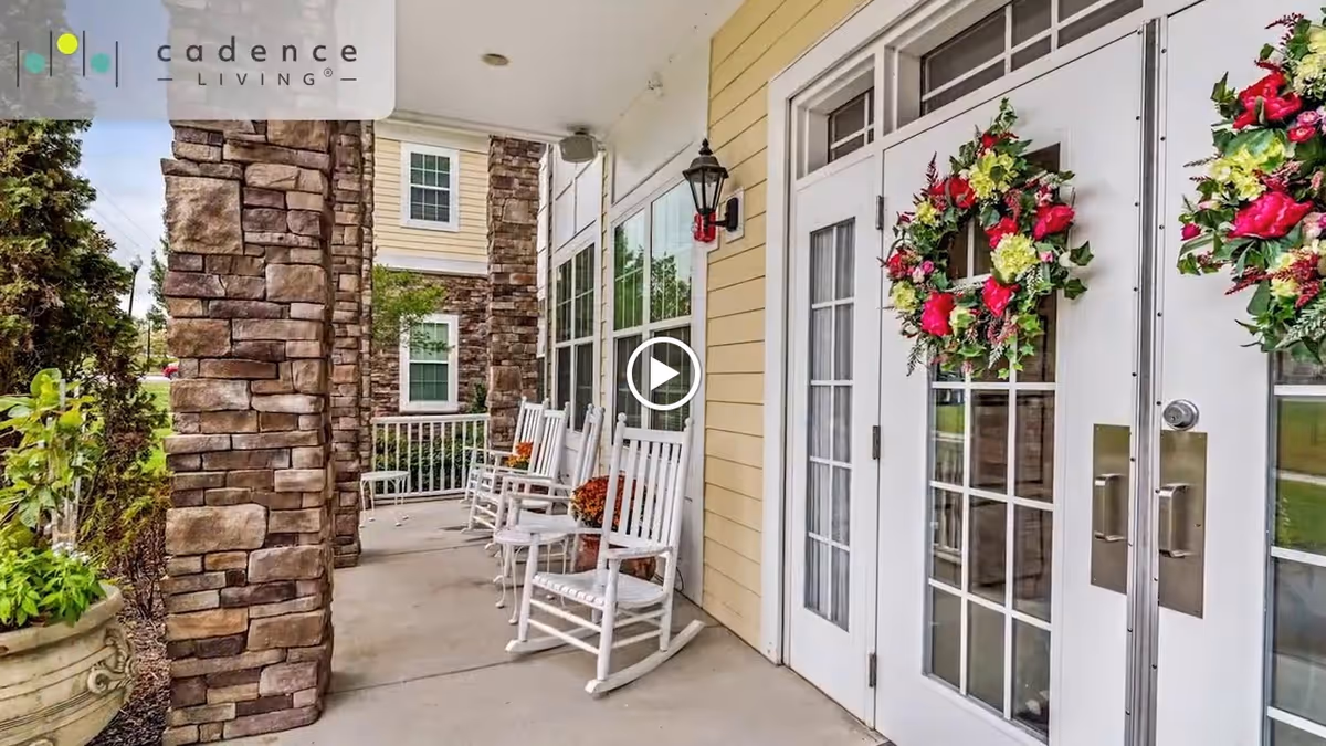 Outdoor covered porch area with white rocking chairs lined up along the wall, stone pillars, potted plants, and double glass doors decorated with floral wreaths.