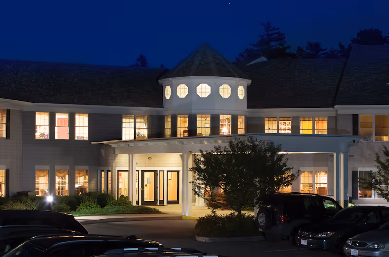 Night view of the front exterior of a senior living facility building with illuminated windows, a covered entrance, and several parked cars in front.