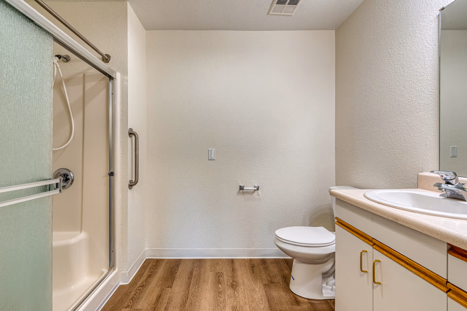 A clean bathroom featuring a shower with sliding glass doors and a grab bar, a toilet, and a sink with a countertop and cabinet below. The floor has wood-like vinyl flooring and the walls are painted light beige.