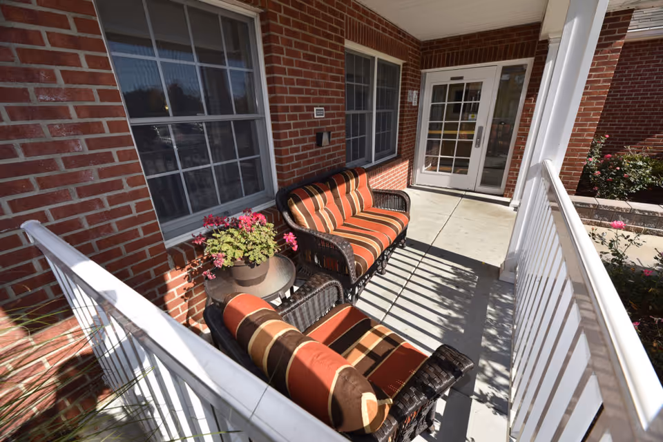 A covered brick-front porch with striped cushioned wicker chairs, a small table with potted flowers, and a glass entrance door.
