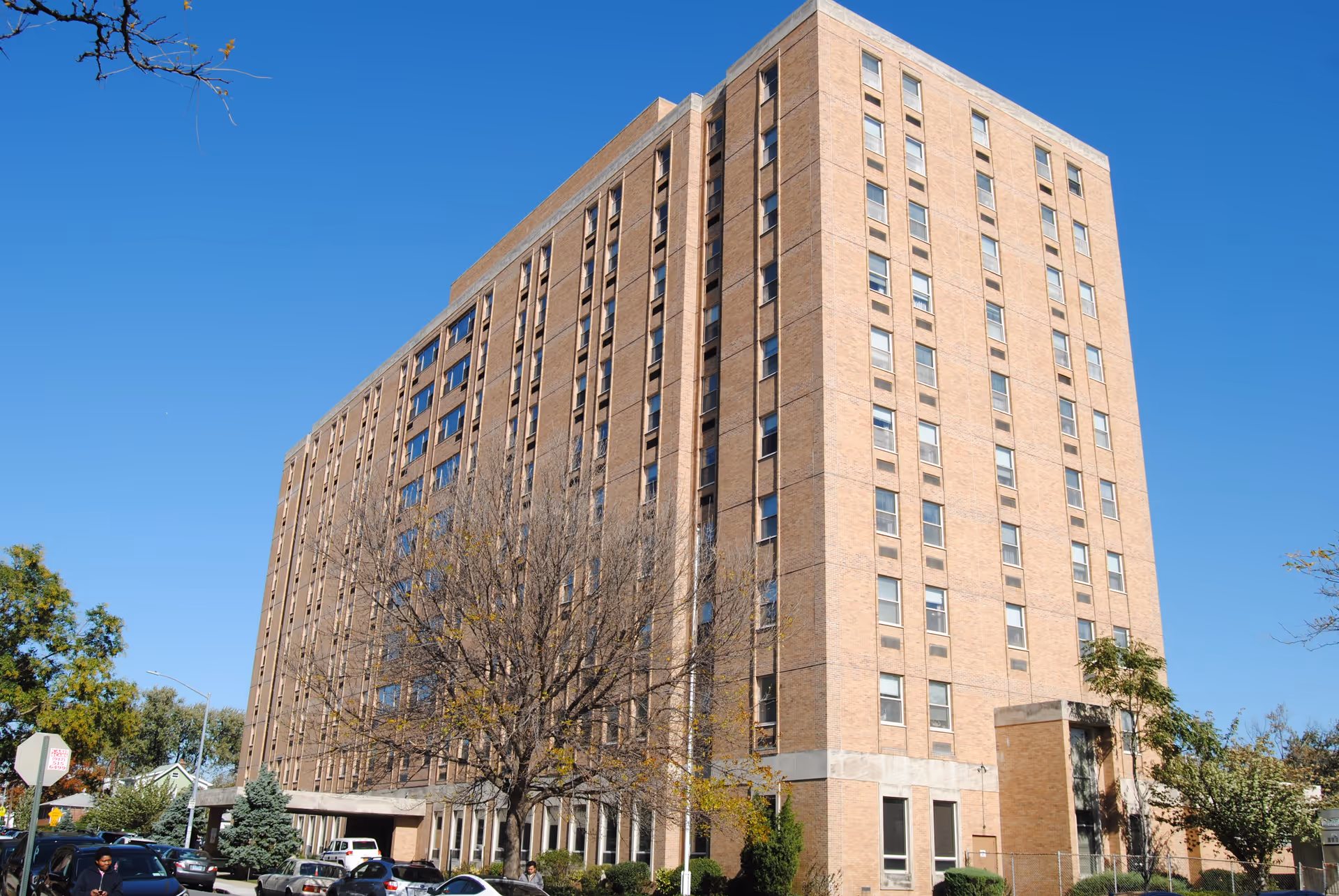 A tall multi-story brick building under a clear blue sky, with several windows and some trees and parked cars in front of it.
