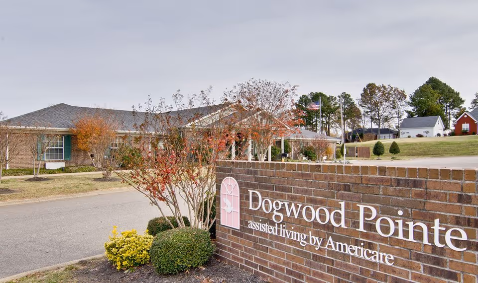 Exterior view of Dogwood Pointe assisted living facility showing a brick sign with the facility name and a single-story building with a gray roof and surrounding trees and bushes.