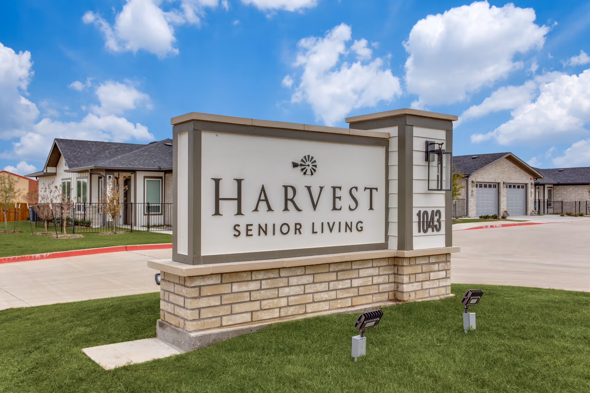 Outdoor view of the entrance sign for Harvest Senior Living with the address number 1043, set on a brick and stone base with a building and driveway in the background under a partly cloudy blue sky.