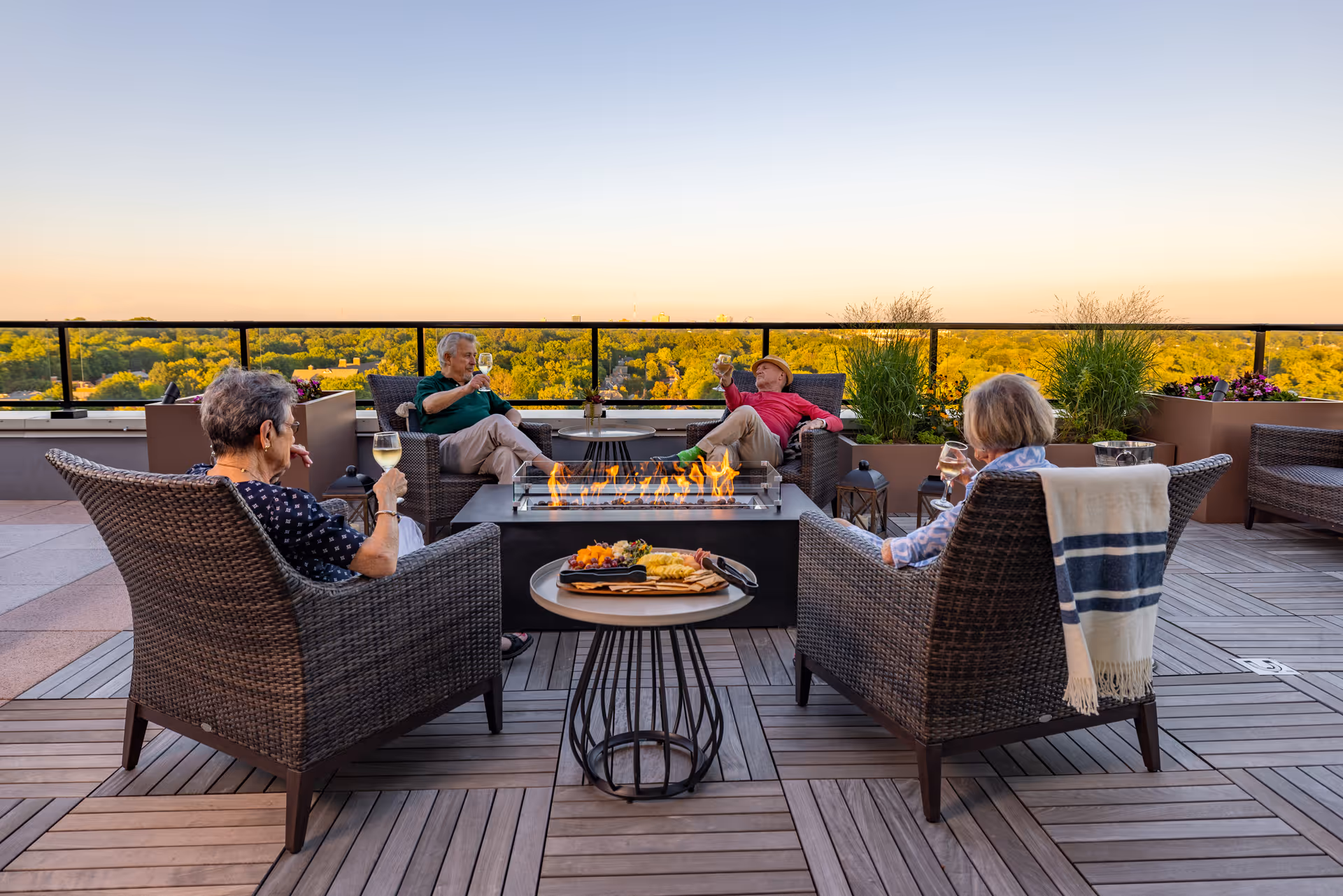Four elderly people sitting in wicker chairs around a rectangular fire pit on a rooftop patio, enjoying drinks and snacks with a scenic view of trees and a clear sky at sunset.