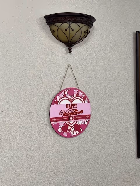 A wall with a decorative wall sconce light fixture mounted near the top and a hanging Valentine's Day decoration below it. The decoration is oval-shaped with a pink and red heart pattern and the text 'Happy Valentine's Day' in the center.