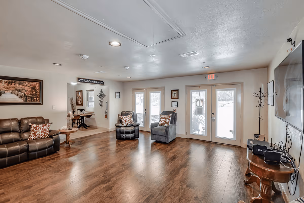 A cozy living room with wooden flooring, two armchairs with patterned cushions, a leather sofa with a matching cushion, a small round wooden table with a lamp, a wall-mounted TV, and double glass doors leading outside. The walls are decorated with framed pictures and a cross, and there is a coat rack near the door.