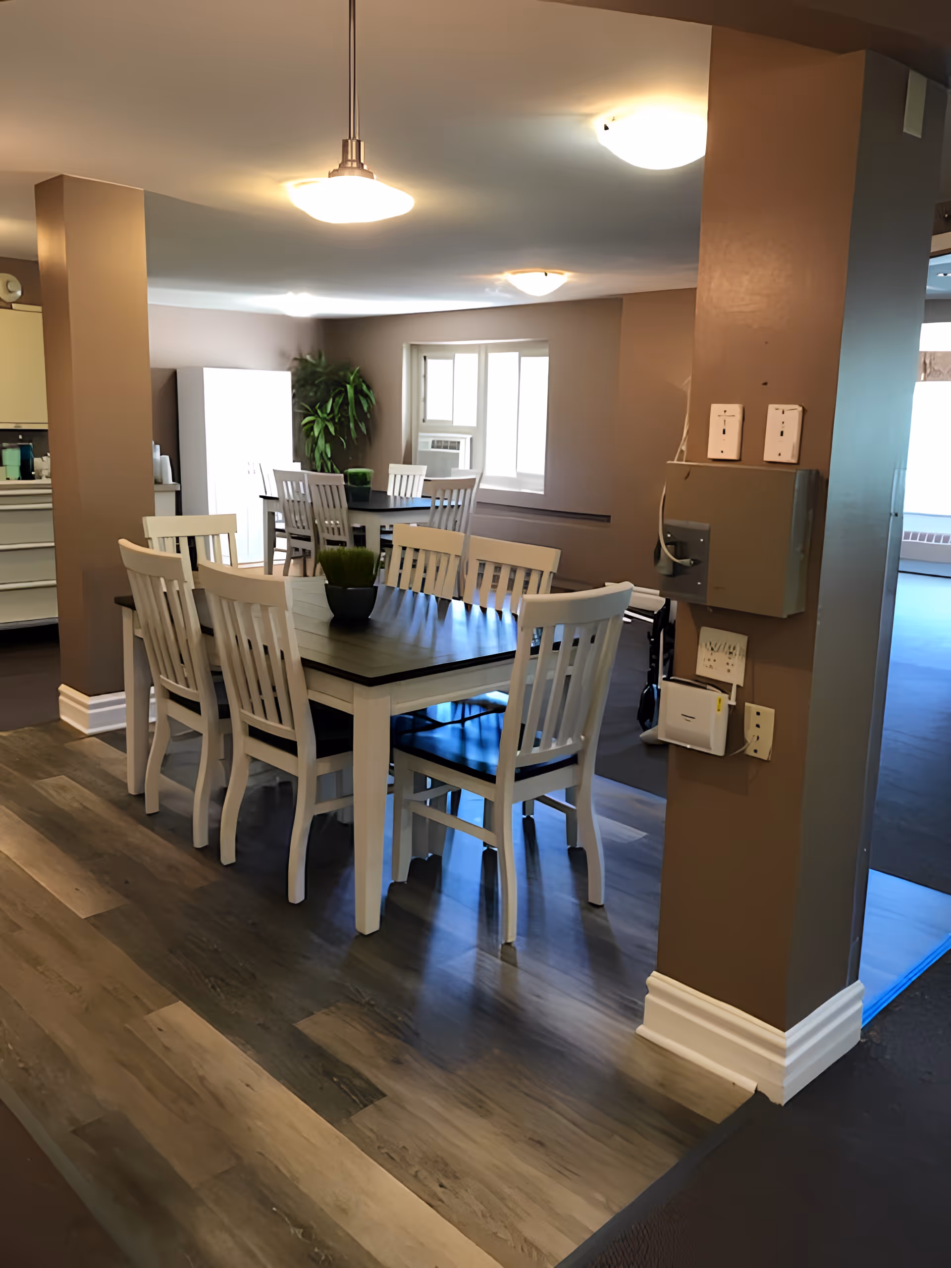 Interior view of a dining area in a senior living facility with two rectangular tables surrounded by white wooden chairs. The room has brown walls, wood-patterned flooring under the tables, and a window with an air conditioning unit. There are ceiling lights and a green potted plant in the corner.