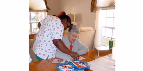 A caregiver assisting an elderly woman seated at a table with art supplies, including markers and paper, in a well-lit room with windows and curtains.