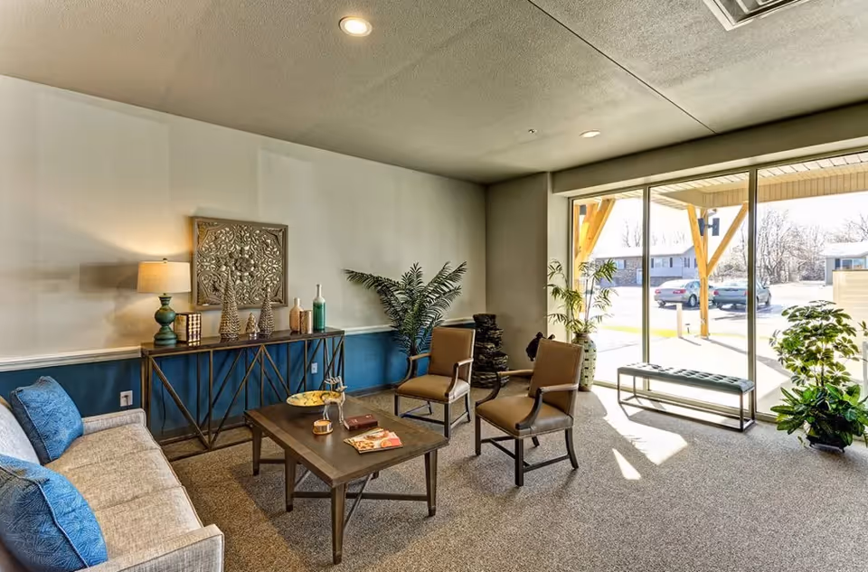 Bright seating area with a sofa, armchairs, coffee table and decorative console beside large floor-to-ceiling windows overlooking the parking area.
