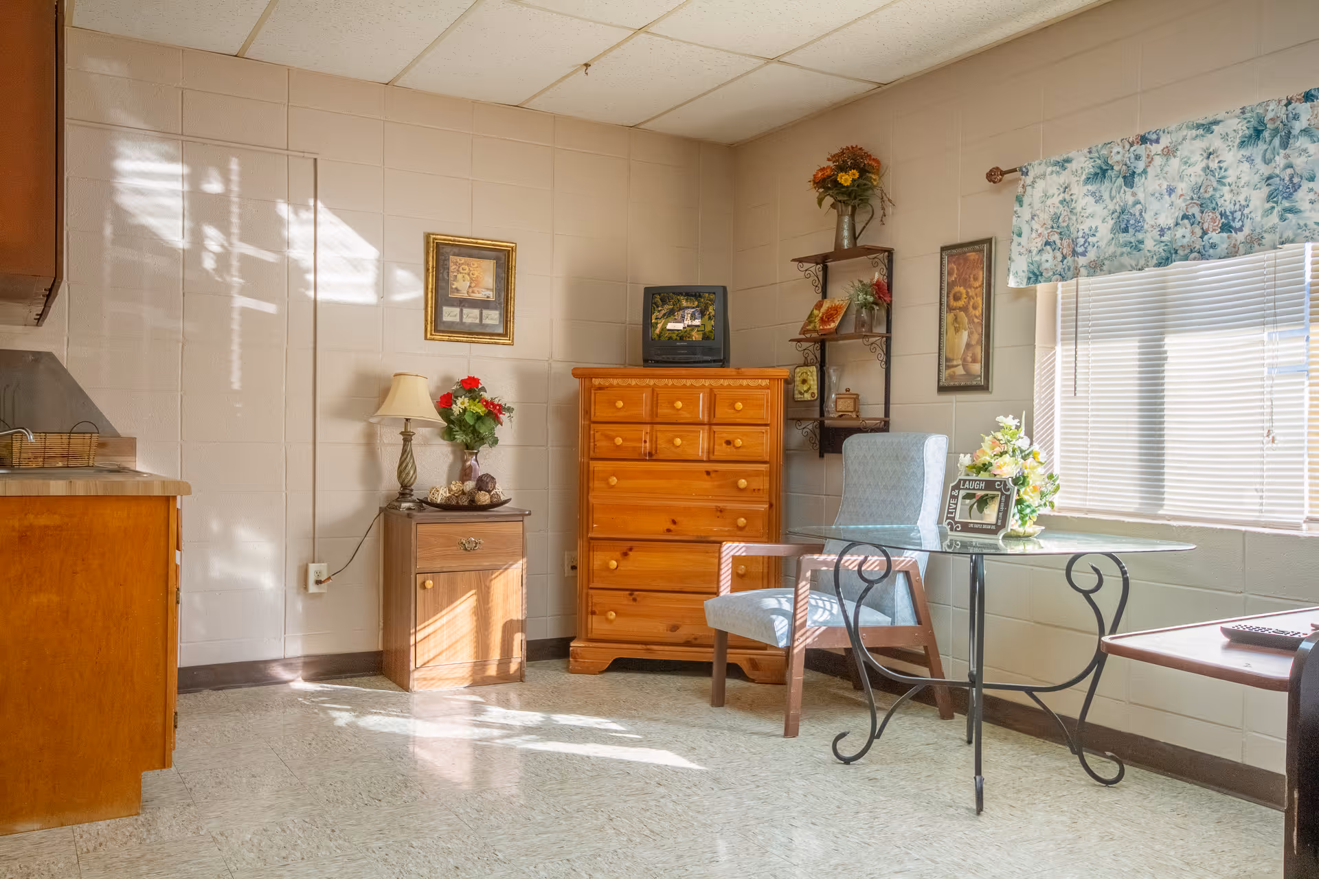 A bright room with a wooden dresser, a small wooden cabinet with a lamp and flowers, a blue upholstered chair, and a round glass table with a flower arrangement and a framed photo. The room has a window with floral curtains and light streaming in, casting shadows on the tiled floor and walls.