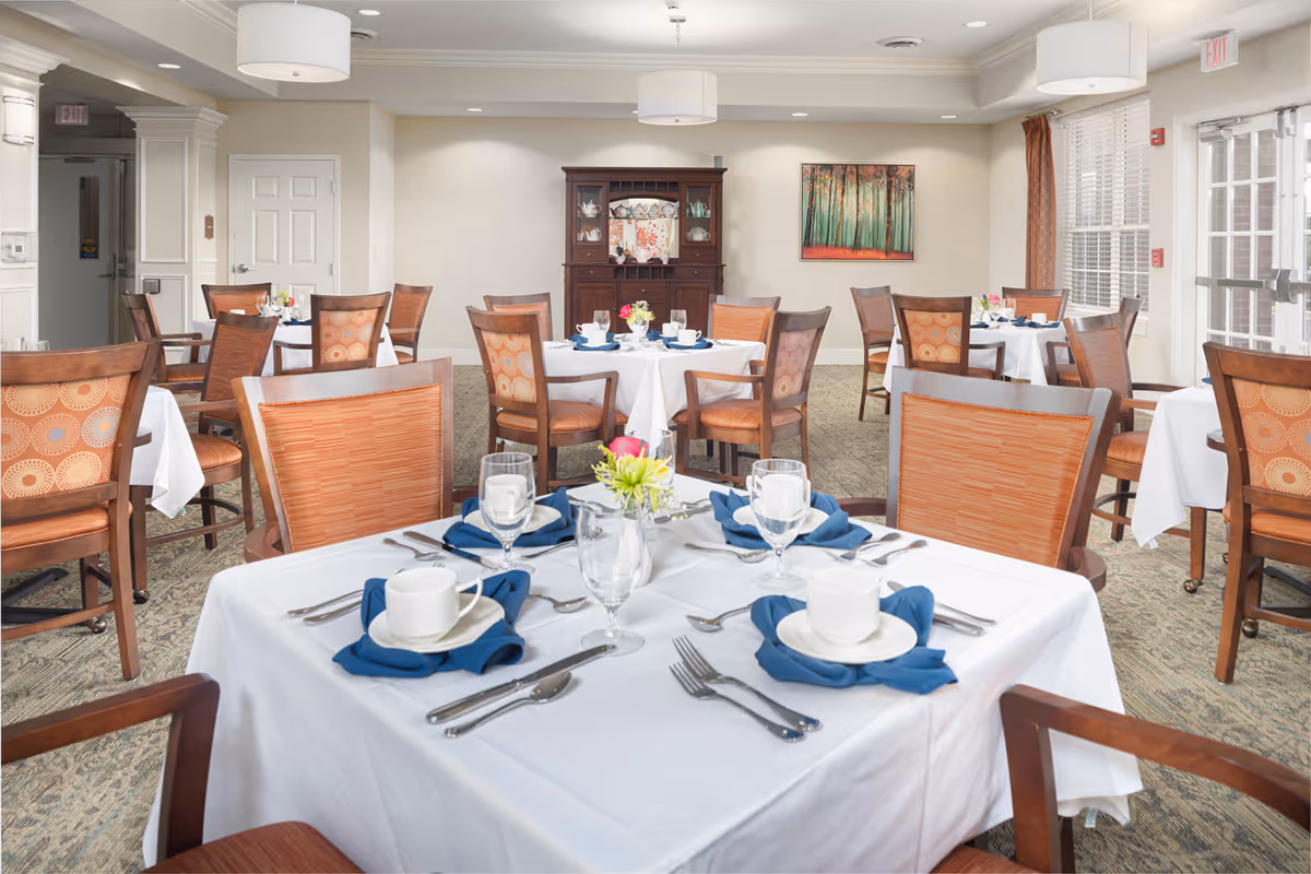 A bright dining room with multiple round and square tables covered with white tablecloths, each set with blue napkins, white cups, glasses, and silverware. Wooden chairs with patterned cushions surround the tables. The room has beige walls, a carpeted floor, a wooden cabinet against the far wall, a painting of a forest, and large windows letting in natural light.