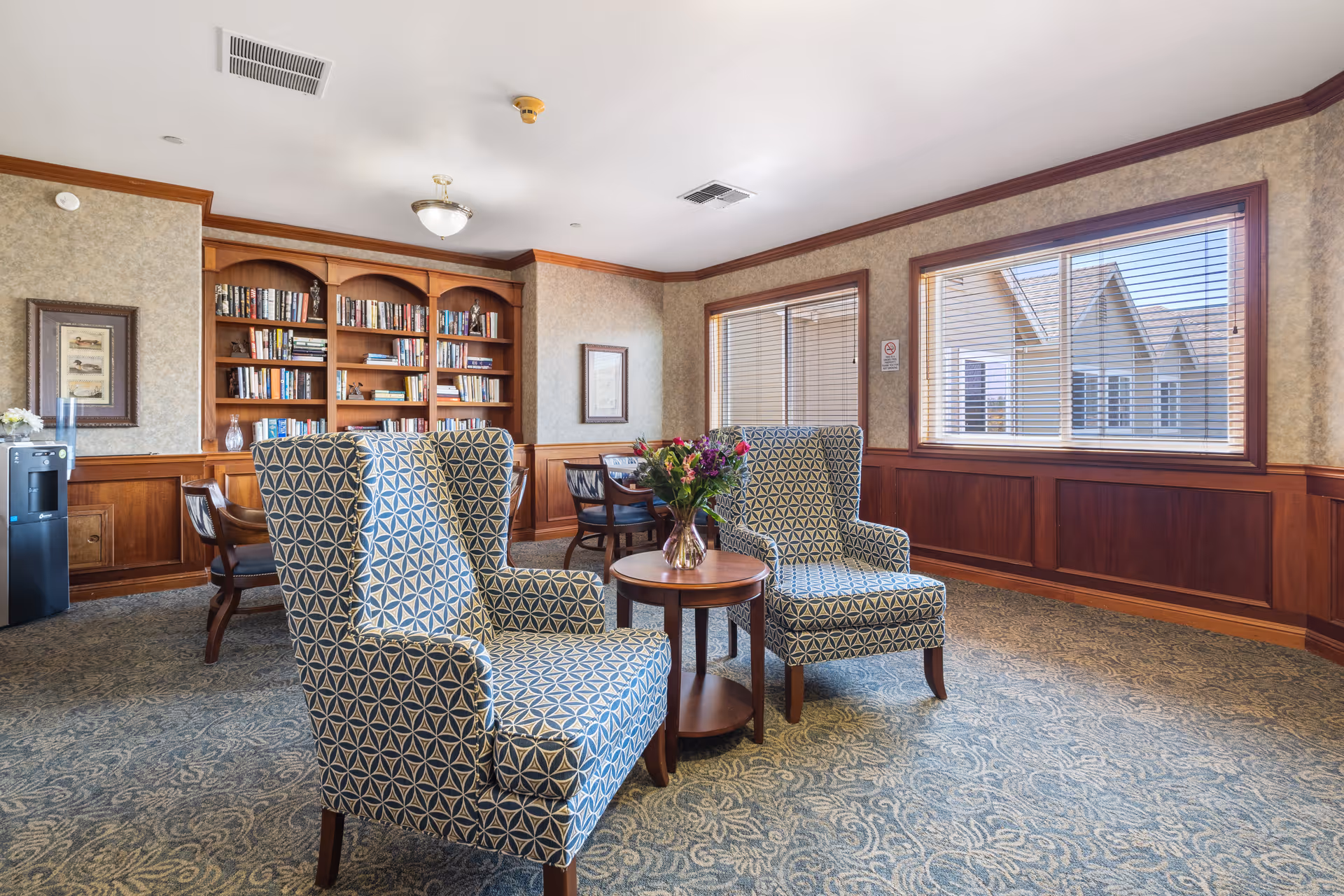 A cozy living room area with two patterned armchairs facing a small round wooden table with a vase of flowers. Behind the chairs is a wooden bookshelf filled with books, framed artwork on the walls, and large windows with blinds letting in natural light. The room features wood paneling and a patterned carpet.