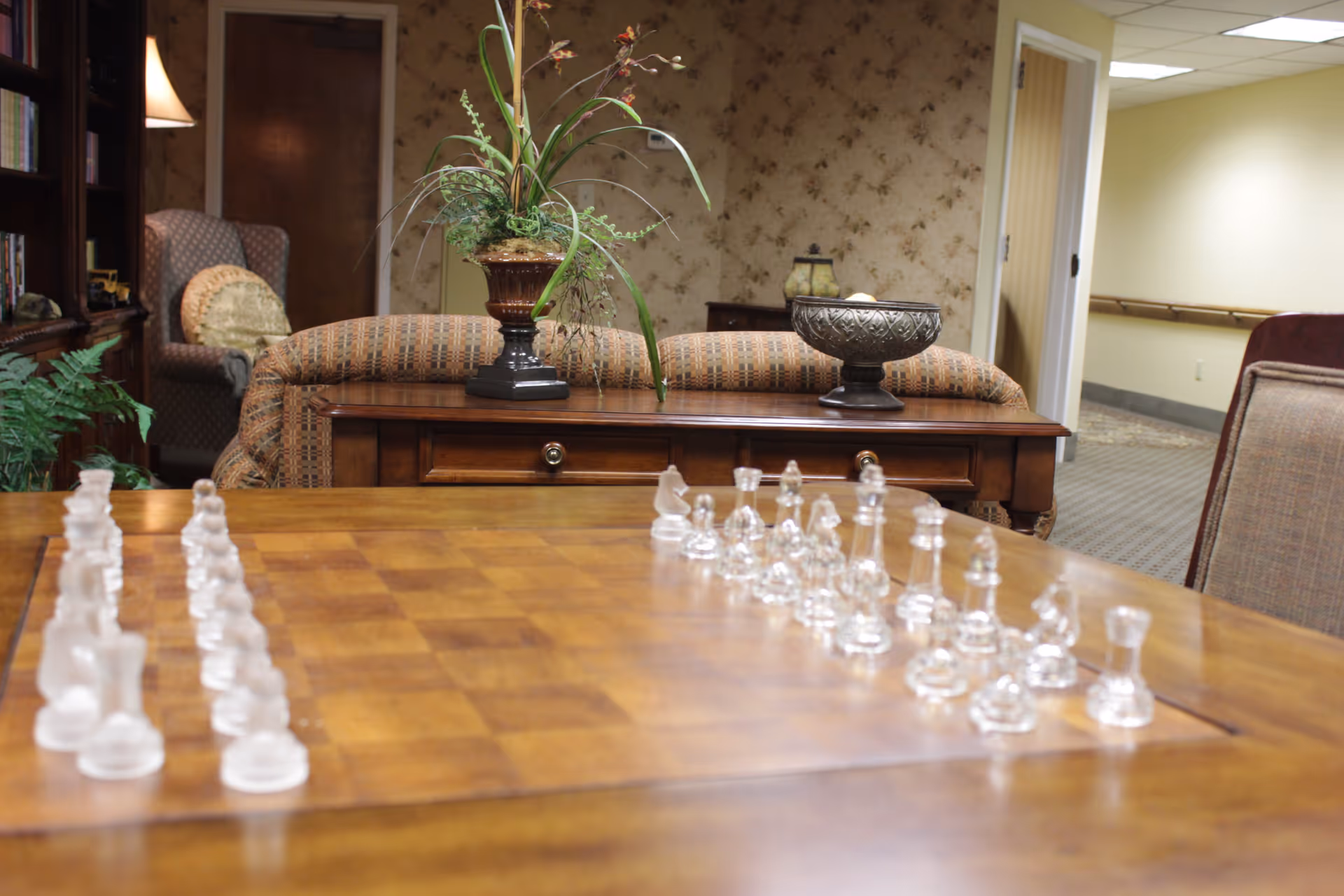 A cozy interior room with a wooden chessboard table set with glass chess pieces in the foreground. Behind the table is a wooden console table with a decorative plant and a bowl. To the left, there is a cushioned armchair with a round pillow and a bookshelf with books and a lamp. The walls have a floral wallpaper and there is a hallway visible to the right.