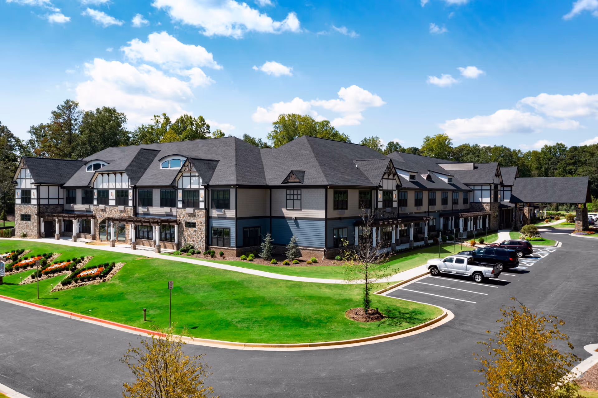 Exterior view of a large two-story senior living facility building with a mix of stone and siding on the facade, surrounded by well-maintained green lawns and flower beds. Several cars are parked in the parking lot adjacent to the building under a partly cloudy blue sky.