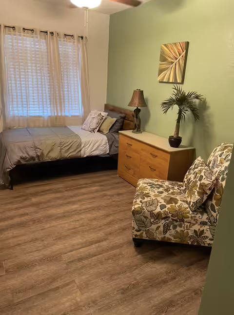 Bedroom with a bed near a window, wooden dresser topped with a lamp and potted plant, and a patterned armchair on wood-look flooring.