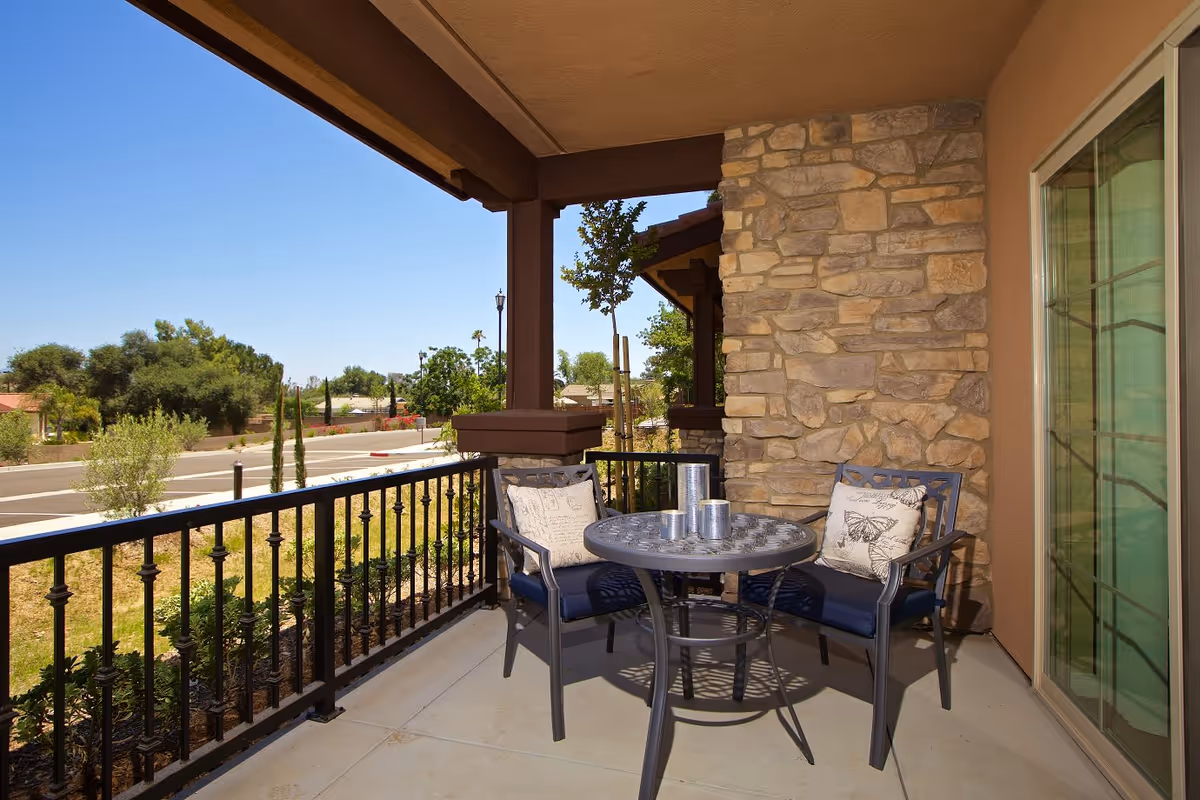 A small outdoor patio area with a round metal table and two chairs with cushions. The patio is enclosed by a black metal railing and has a stone wall on one side. There are three silver candle holders on the table. The patio overlooks a street and some greenery with trees and shrubs under a clear blue sky.