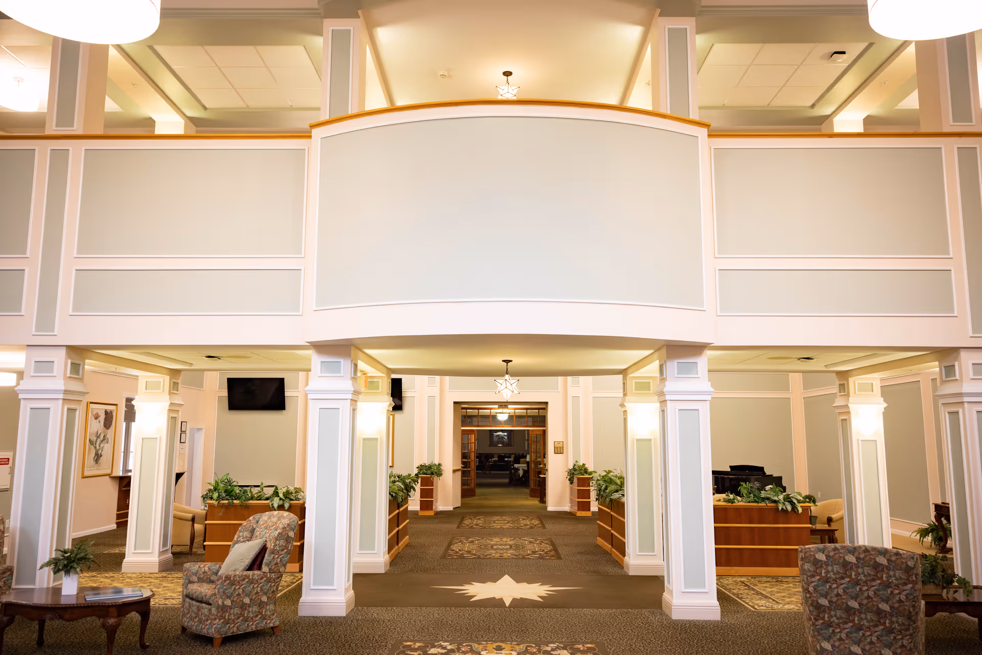 Spacious and well-lit interior common area of a senior living facility with high ceilings, decorative columns, patterned armchairs, wooden tables, plants, and a carpeted floor featuring a star design in the center.