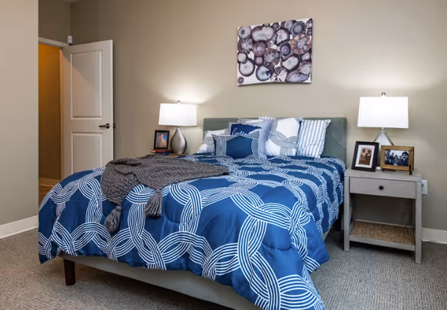 Well-lit bedroom with a blue-patterned bed, two bedside tables with lamps and framed photos, and artwork above the headboard.