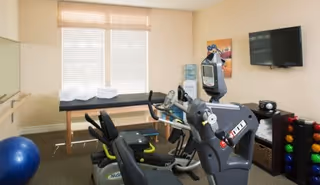 Small fitness room with stationary bikes and cardio machines, a rack of colorful dumbbells, an exercise ball, a wall-mounted TV, and a bench beneath a window.
