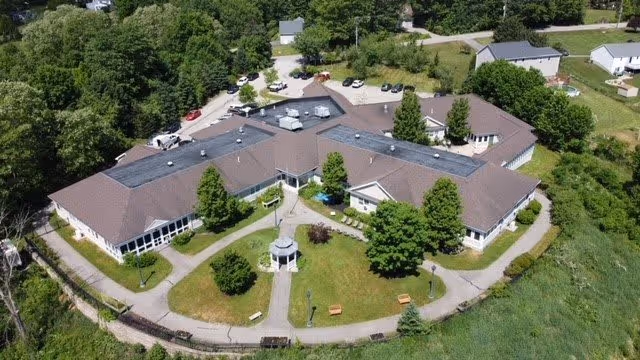 Aerial view of Watson Fields senior living facility showing a large, single-story building with a brown roof surrounded by green trees and grass. The building is arranged in a U-shape with pathways, benches, and a small gazebo in the courtyard area. Several cars are parked near the entrance, and the facility is located in a semi-rural area with other buildings and greenery nearby.
