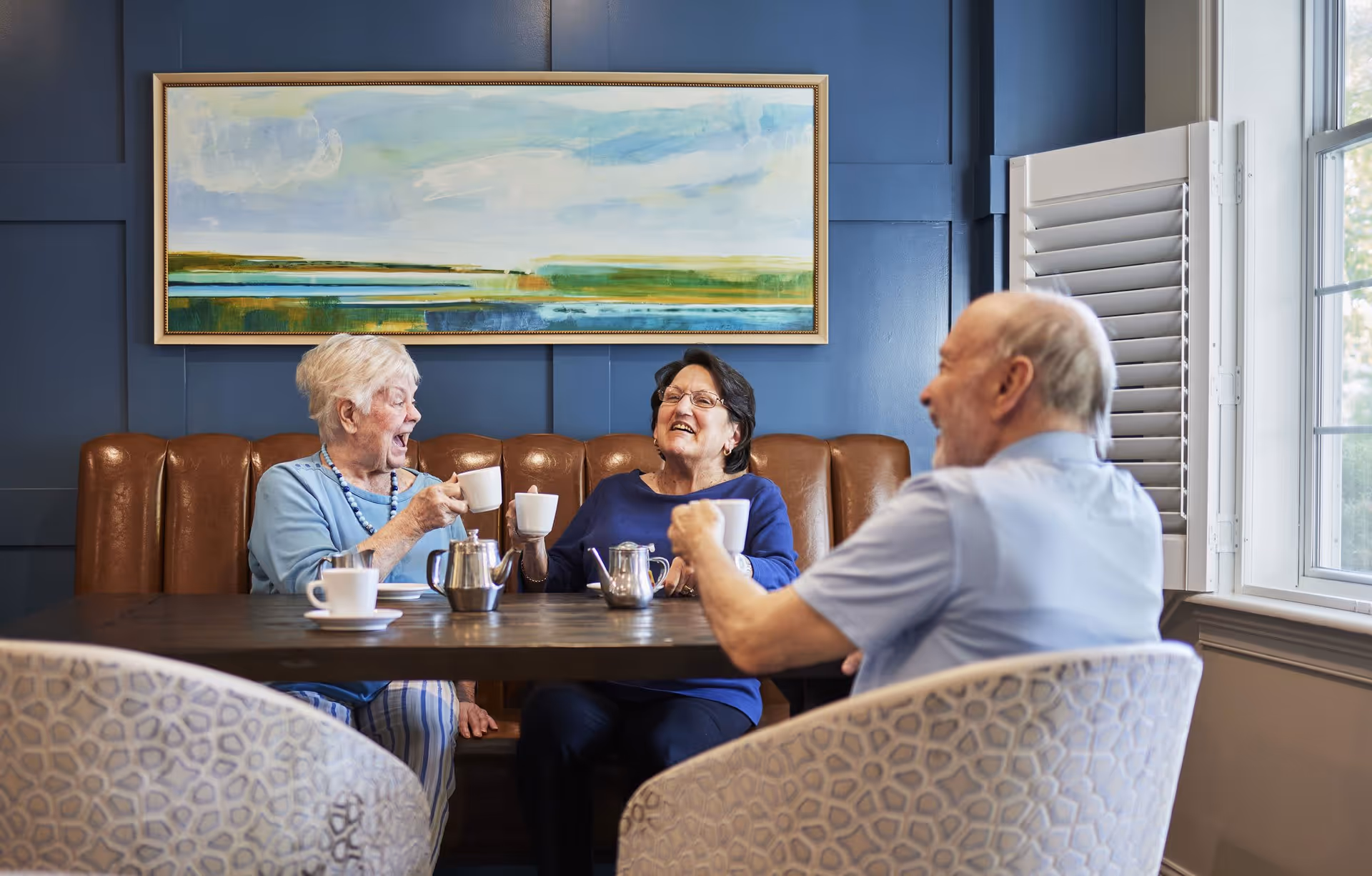 Three elderly people sitting around a table in a cozy room with blue walls and a large window. Two women and one man are smiling and holding white coffee cups, enjoying a social moment. A landscape painting hangs on the wall behind them.