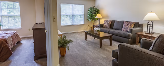 A bright and spacious senior living unit featuring a living room and a bedroom separated by a partial wall. The living room has a gray sofa with floral pillows, a matching armchair, a wooden coffee table with a small plant, two side tables with lamps, and a large window letting in natural light. The bedroom has a bed with a brown comforter, a wooden dresser, and a window with blinds.