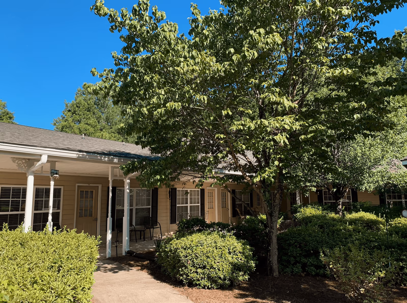 Exterior view of a senior living facility building with beige siding, multiple doors and windows, a covered porch with white columns, surrounded by green bushes and trees under a clear blue sky.