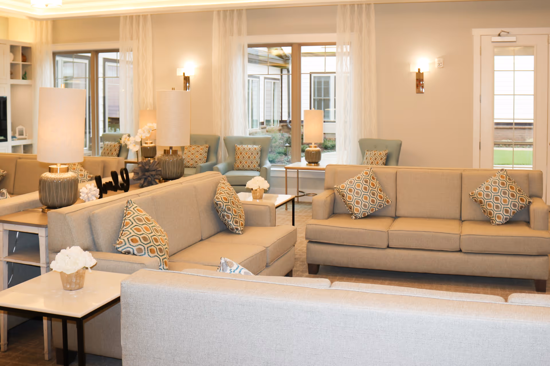 A bright and cozy living room with beige sofas adorned with patterned cushions, light blue armchairs, side tables with lamps, and decorative flowers. Large windows with sheer white curtains allow natural light to fill the space.