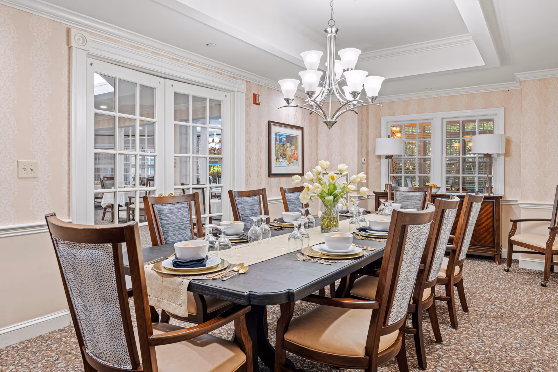 A well-lit dining room with a long dark wooden table set for eight people. The table is decorated with white bowls, plates, glasses, gold-colored cutlery, and a vase of white tulips in the center. The room features patterned wallpaper, a chandelier overhead, and large windows with white frames. There are wooden chairs with cushioned seats around the table, and a sideboard with two lamps and a framed picture on the wall.
