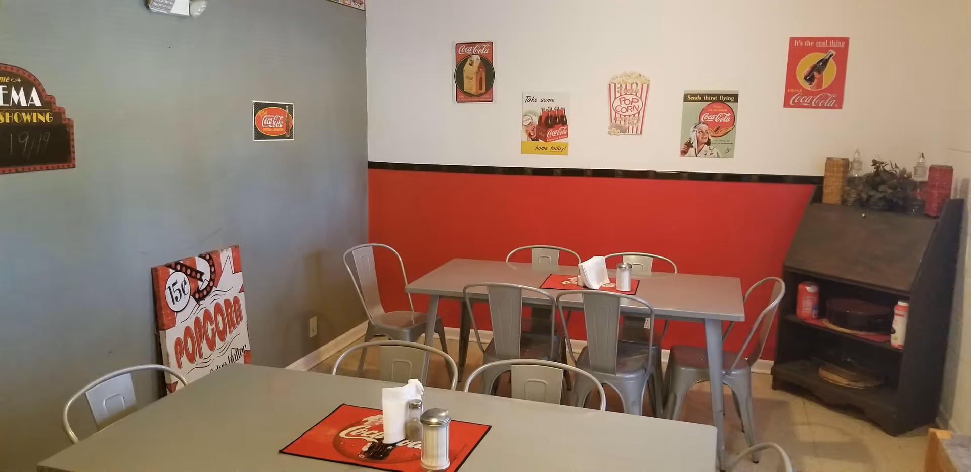 A small dining area with two gray tables and metal chairs. The walls are painted gray and red with vintage Coca-Cola and popcorn-themed signs decorating the walls. There is a black shelving unit in the corner holding various items.