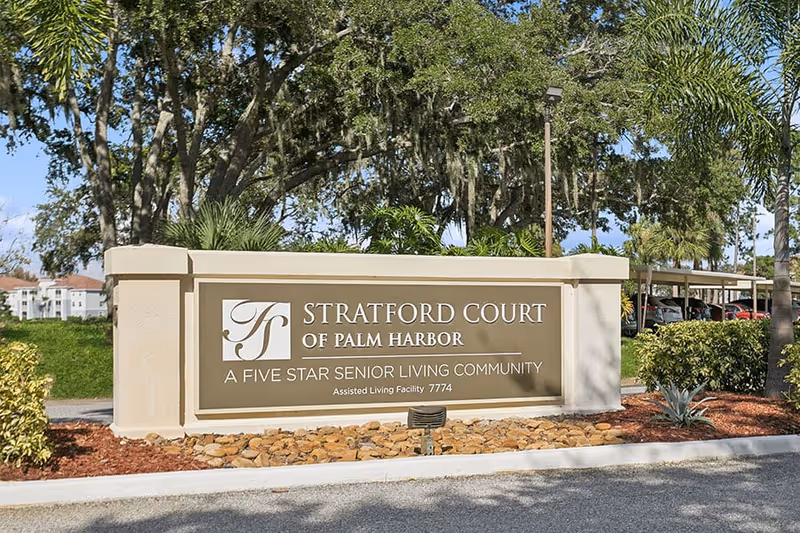 Entrance sign for Stratford Court of Palm Harbor, a five star senior living community, surrounded by landscaping with trees and plants in the background.