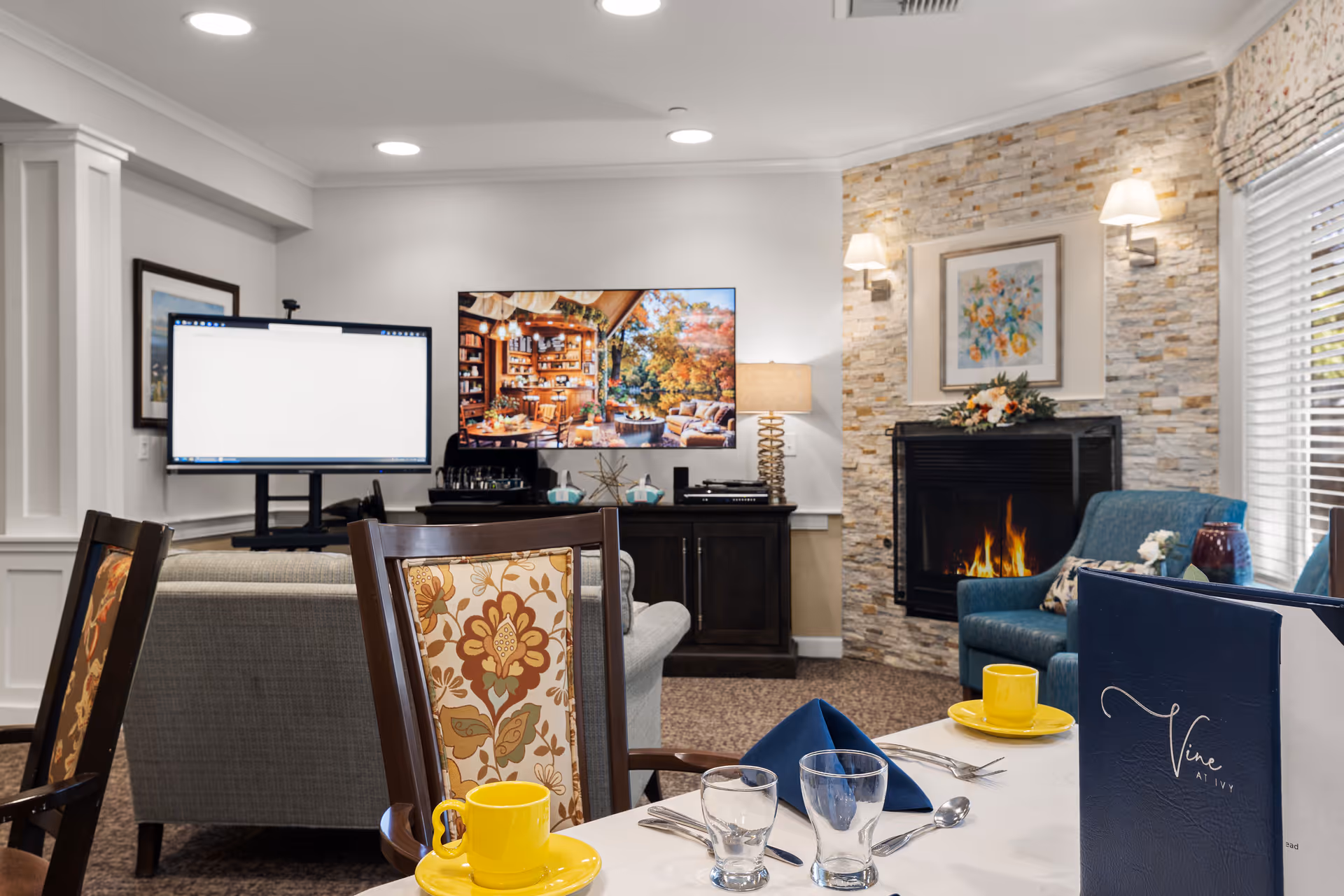 A cozy living room area with a stone fireplace lit with a fire, a blue armchair, a gray sofa, and a TV mounted on a dark wooden cabinet. In the foreground, a dining table is set with yellow cups and saucers, glasses, silverware, and a blue napkin. A menu with the text 'Vine at Ivy' is visible on the table. The room has soft lighting from wall sconces and a table lamp, with a framed floral painting above the fireplace.