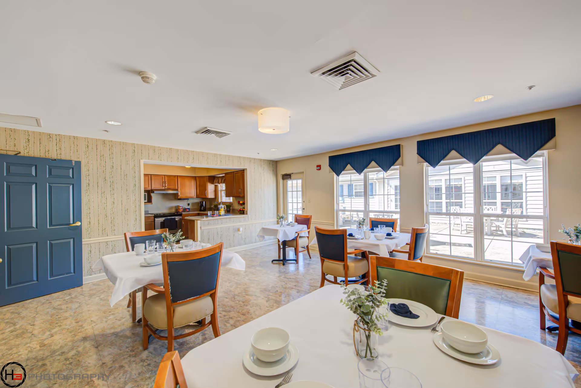 Bright dining room with several set tables and chairs, large windows, and an open serving area to a kitchen.